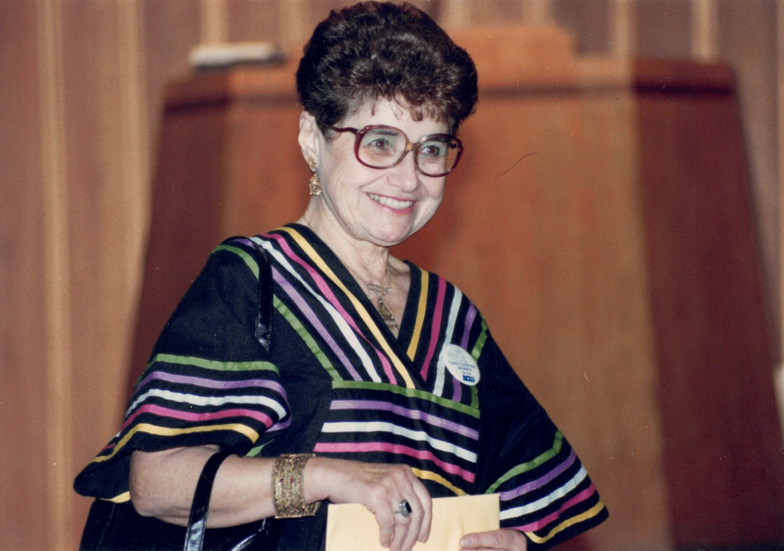 Hedy Epstein smiling in front of a podium wearing a black dress with colorful ribbons on the neckline and sleeves