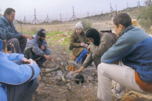 Hedy Epstein in a brown winter coat and gray woolen cap sitting around a fire with Palestinian farmers. The separation fence is in the background. The farmers are waiting to see if the gate will be opened by Israeli soldiers so they can access their agricultural fields.
