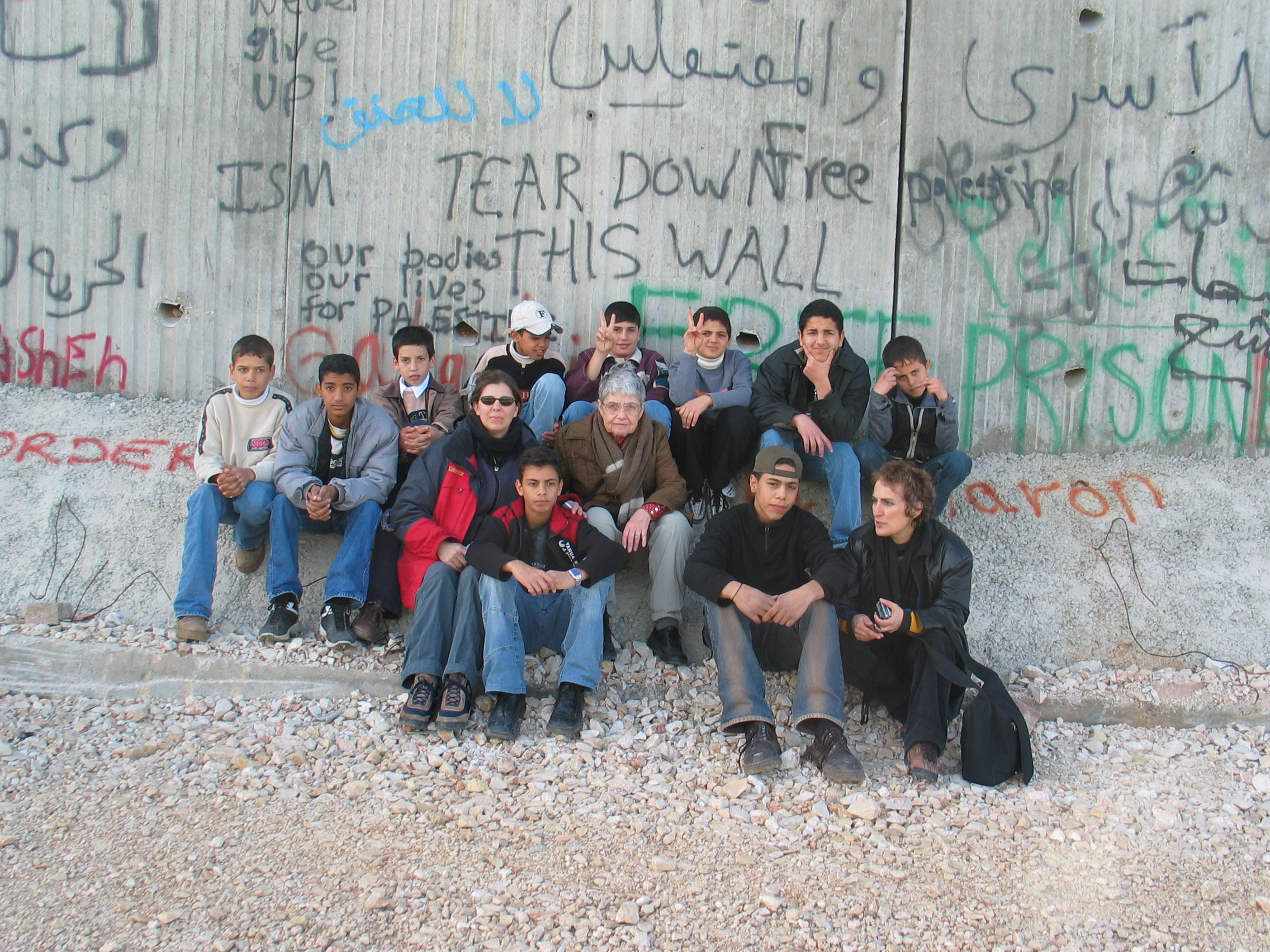 Hedy Epstein seated with a group of boys and young men and her travel companions in front of the 25' concrete separation wall in Qalqilya, West Bank