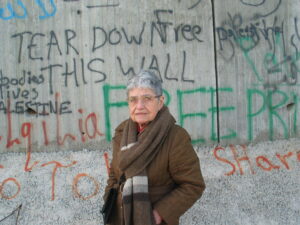 Hedy in a brown winter coat and scarf with hands in her pockets standing in front of the 25' concrete separation wall in Qalqilya, West Bank. The wall is covered with graffiti, Tear Down This Wall, Free Palestine.