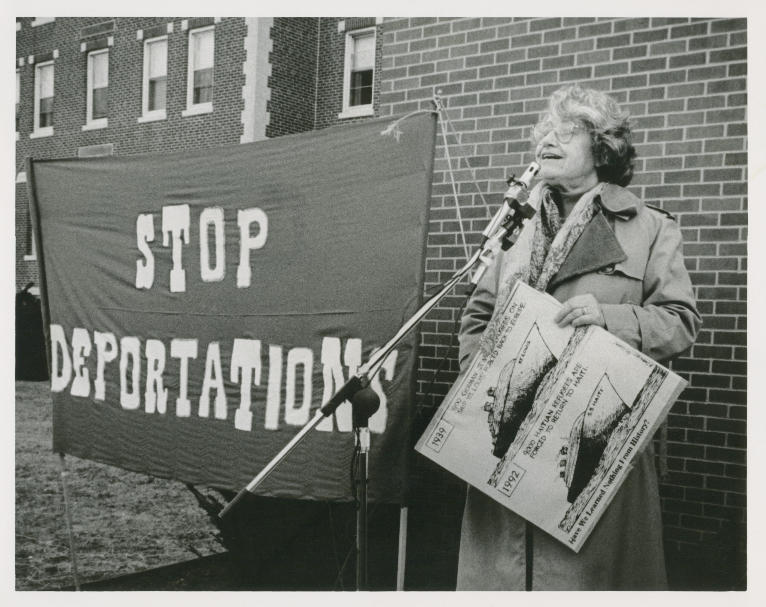 Hedy Epstein is standing at a microphone next to a large banner that says stop deportations. She is carrying a sign showing two ships, one is the 1939 ship St. Louis, the other ship is deporting Haitian refugees