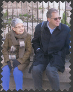 Hedy and Mark are wearing winter coats and are seated in front of a gate with a stone wall behind them
