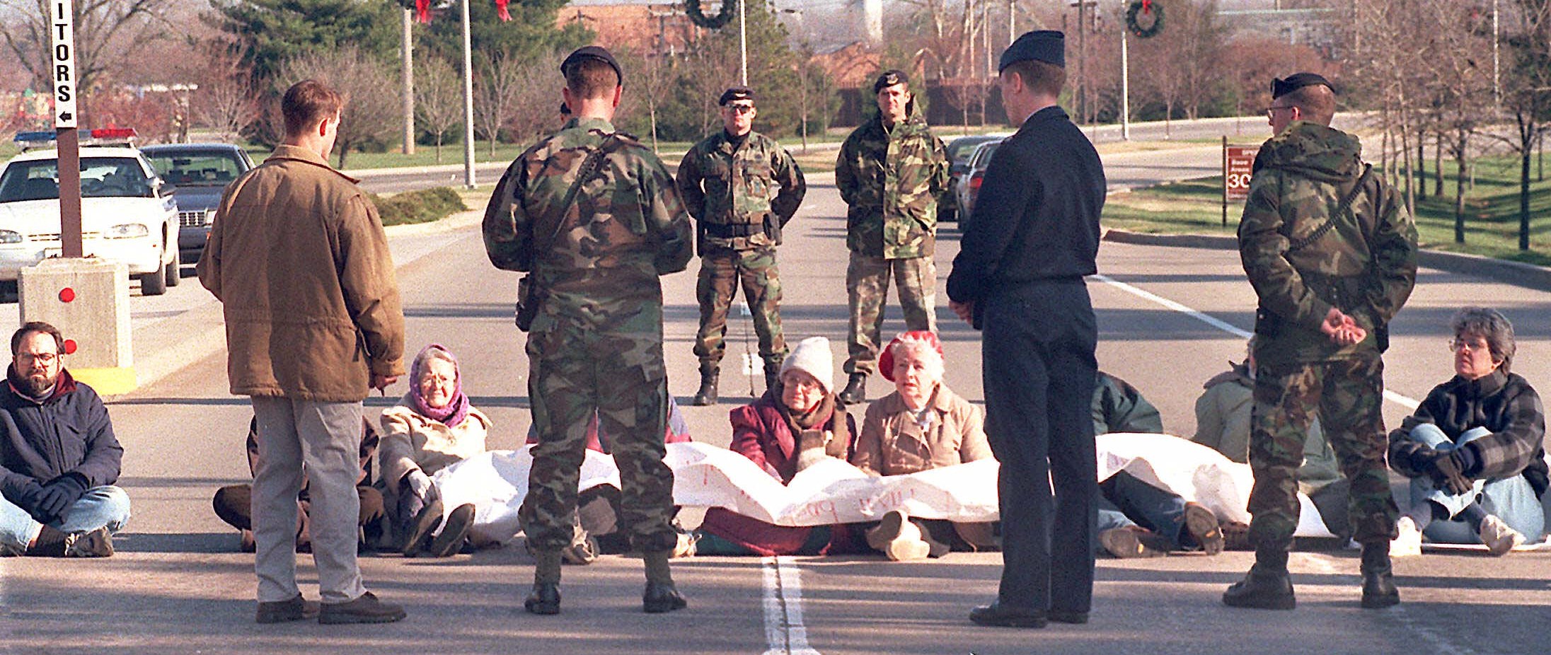 Hedy Epstein and several protesters opposed to the U.S. bombing of Iraq are seated on the road blocking the gate to Scott Air Force Base. Military personnel stand in front and behind the protesters.