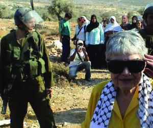 Hedy Epstein standing in Bil'in, a village in the West Bank. An Israeli soldier is to her side and another is behind her. Palestinian women from the village are behind the soldiers.
