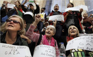 Hedy Epstein seated between two women with a crowd behind, fist raised, wearing a sign that says "Hunger Strike for Gaza."