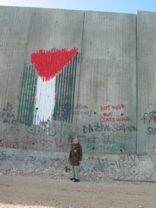 Hedy Epstein standing in front of the 25' concrete separation wall in Qalqilya West Bank. Hedy looks very small compared to the full height of the wall. There is graffiti and a large Palestinian flag painted on the wall.