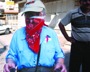 Hedy Epstein in a gray baseball cap with WCA printed in red, a blue button up shirt, and a red bandana across her nose and mouth. She is holding a spent tear gas container.