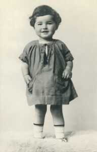 A smiling 3 year old Hedy Epstein with short dark hair, wearing a dress, standing up, looking straight ahead