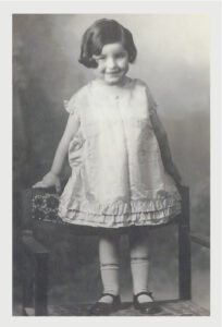 Black and white photo of a child, she is standing on a chair, smiling, wearing a dress and black buckle shoes.