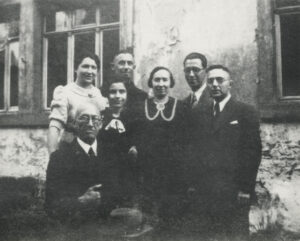 Hedy's grandfather is seated, wearing a suit holding his hat in his lap, standing behind him are 13 year old granddaughter Hedy Epstein and his three adult children, a daughter-in-law and son-in-law
