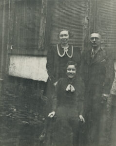 Black and white photo of a couple standing behind their seated 12 year old daughter, they are outside standing in front of the back of a building
