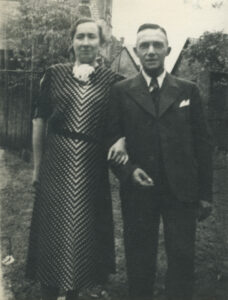 Black and white photo of a woman and man standing outside in a yard. Her arm is intertwined with his arm. She is wearing a dress, he is wearing a suit and tie.