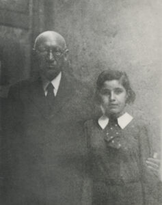 Black and white photo of an elderly gentleman standing with his arm around a young girl, he is wearing a dark suit, she is wearing a dark dress with a white collar and a polka-dot bow, their expressions are solemn