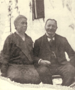 Black and white photo, couple is seated side by side, they are outdoors on a bench in front of a small window.