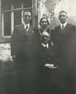 An elderly gentleman seated with hands folded in front of his adult children; the three men are wearing suits and ties, the woman standing in the middle is wearing a dress
