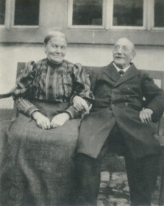Black and white photo of an elderly couple seated outside on a bench, both smiling, she is wearing a plaid blouse and a dark floor-length skirt, her hands are resting in her lap, he is wearing a dark overcoat with his arm intertwined with hers.