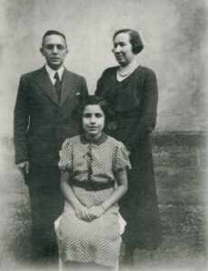 Black and white photo of parents standing behind their seated 12 year old daughter, the father is wearing a dark suit, the mother is wearing a dark dress, the daughter is wearing a printed dress.