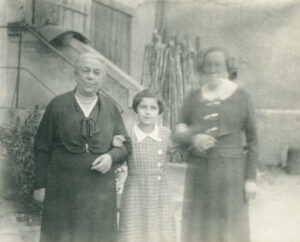 Elderly woman on the left, twelve year old girl in the middle and her mother to the right. They are standing in front of an external stair with their arms intertwined.