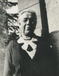 Hedy Epstein's grandmother, an elderly woman standing outside next to a building with a large evergreen tree behind her