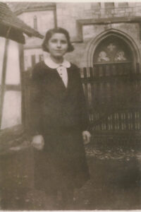 Black and white photo of a 14 year old girl dressed in a white blouse, dark cardigan and skirt, standing in a yard next to a church with a stained glass window